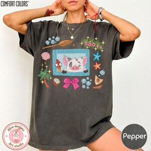 Puede incluir: Camiseta Comfort Colors color pimienta con una ilustraci&oacute;n de dibujos animados de un cerdo y un gallo en un marco azul, rodeada de elementos tropicales. La camiseta tambi&eacute;n tiene el texto "Alice designz art studio".