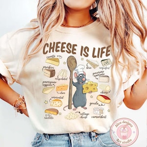 Puede incluir: Camiseta color crema con el texto "CHEESE IS LIFE" e ilustraciones de varios quesos. Un chef de dibujos animados con forma de rata sostiene una cuchara de madera y un bloque de queso. Los tipos de queso incluyen Comte, Brie, Gouda, y m&aacute;s.