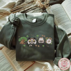 May include: Green crewneck sweatshirt with a cartoon safari design. The design includes a tree, a character with binoculars, a jeep, and a compass. Paw prints and foliage are also included in the design.