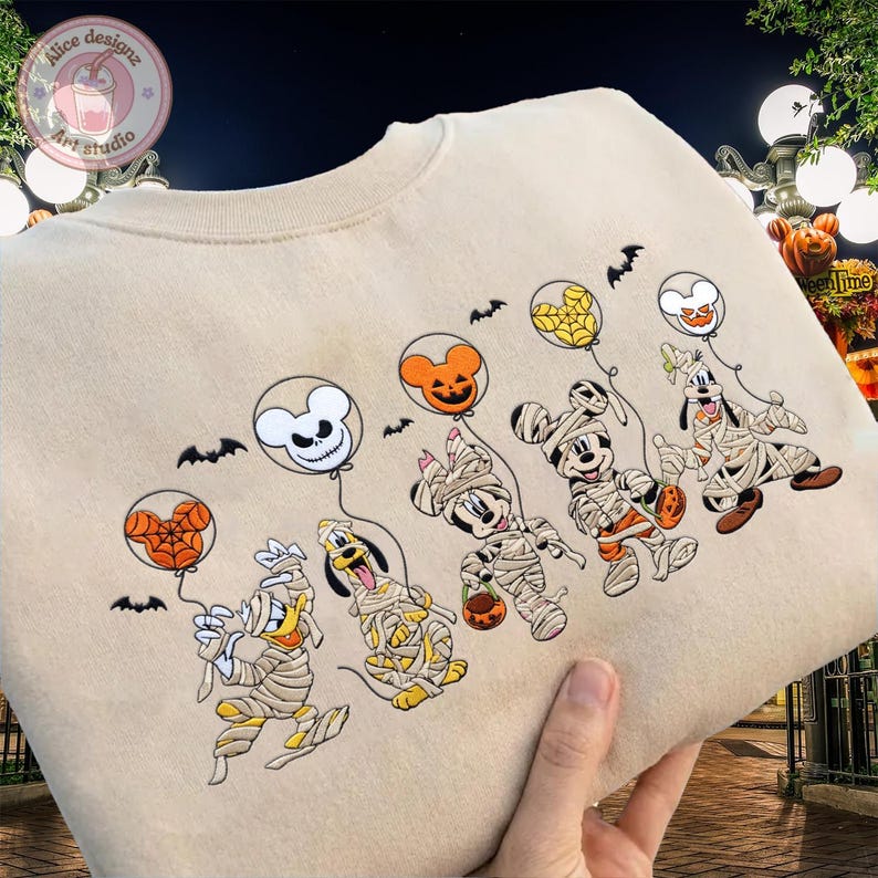 May include: Beige sweatshirt featuring a Halloween-themed design. The design includes cartoon characters dressed as mummies, holding balloons with jack-o'-lantern and skull designs. Black bats are scattered around the characters. The sweatshirt has a crew neck.