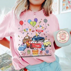 Op de afbeelding: Roze T-shirt met een kleurrijk verjaardagsontwerp met cartoonauto's, ballonnen en een taart. Het ontwerp bevat het nummer 3, een banner met de tekst "BIRTHDAY" en een ruimte voor een naam. Een mok met de tekst "Alice designz Art studio" is ook zichtbaar.