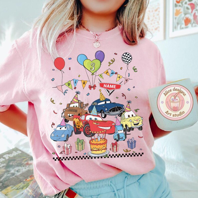 Op de afbeelding: Roze T-shirt met een kleurrijk verjaardagsontwerp met cartoonauto's, ballonnen en een taart. Het ontwerp bevat het nummer 3, een banner met de tekst "BIRTHDAY" en een ruimte voor een naam. Een mok met de tekst "Alice designz Art studio" is ook zichtbaar.