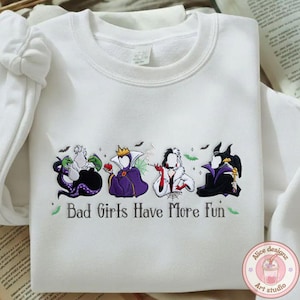 May include: White sweatshirt featuring embroidered cartoon villains with the text "Bad Girls Have More Fun." The design includes Ursula, the Evil Queen, Cruella de Vil, and Maleficent. The design is in shades of purple, black, white, and green.