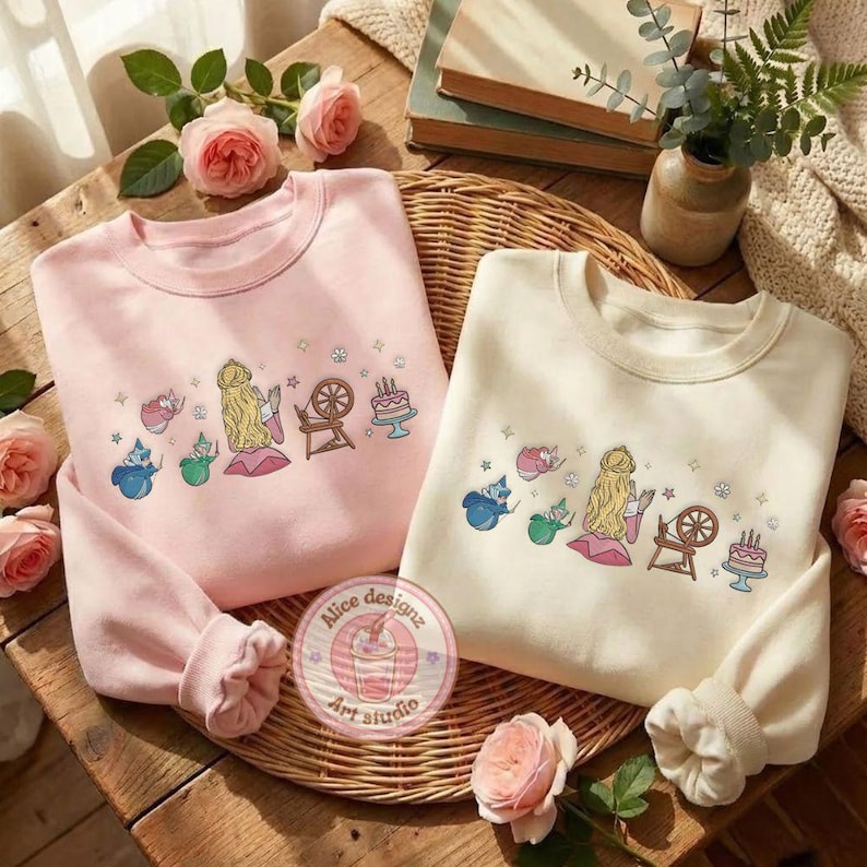 May include: Two crewneck sweatshirts, one light pink and one cream, with embroidered fairytale designs. The designs include a princess, fairies, a spinning wheel, and a cake. The sweatshirts are displayed on a wooden surface with roses.