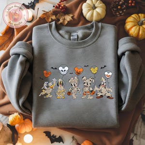 May include: A gray sweatshirt featuring a Halloween-themed graphic. The design includes cartoon characters dressed as mummies, holding pumpkin-shaped treat buckets and balloons. The balloons are in the shape of Mickey Mouse, Minnie Mouse, and other characters.