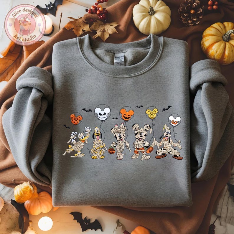 May include: A gray sweatshirt featuring a Halloween-themed graphic. The design includes cartoon characters dressed as mummies, holding pumpkin-shaped treat buckets and balloons. The balloons are in the shape of Mickey Mouse, Minnie Mouse, and other characters.