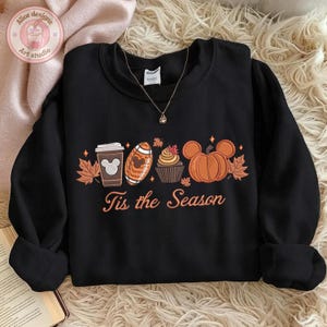 May include: Black crewneck sweatshirt with a fall-themed graphic. The design includes a coffee cup, football, cupcake, and pumpkin, all with Mickey Mouse ears, along with the text "Tis the Season".