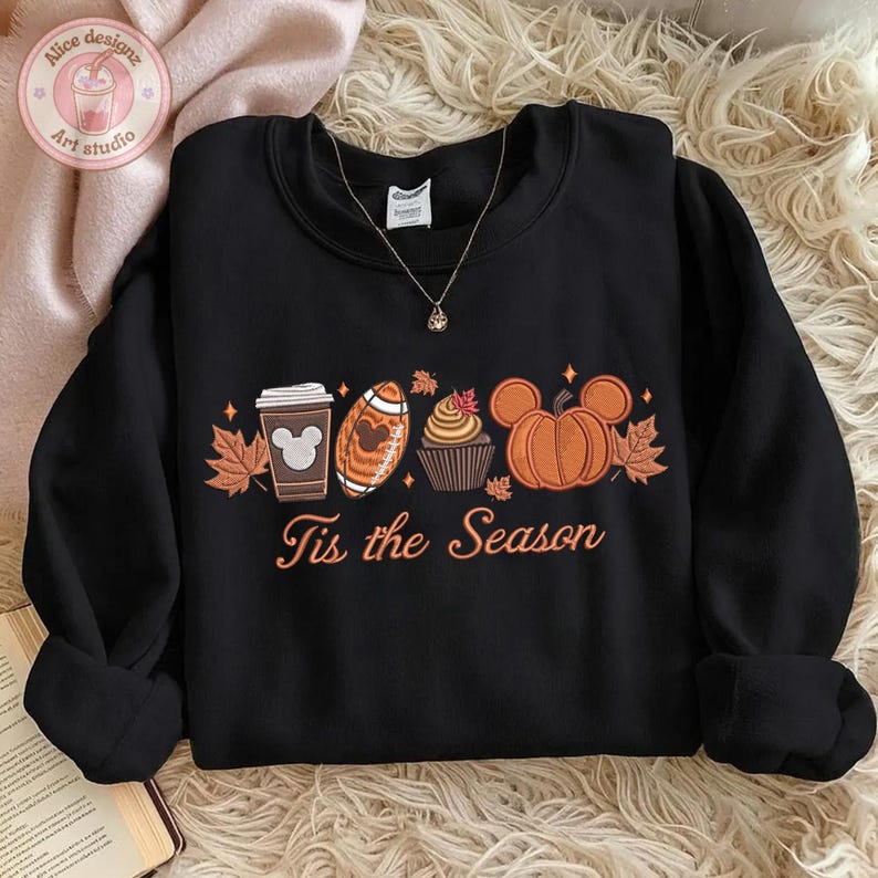 May include: Black crewneck sweatshirt with a fall-themed graphic. The design includes a coffee cup, football, cupcake, and pumpkin, all with Mickey Mouse ears, along with the text "Tis the Season".