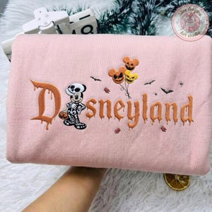 May include: A light pink sweatshirt featuring embroidered orange text that reads Disneyland in a dripping font. The design includes a skeleton-costumed mouse character, three jack-o'-lantern balloons, and small black bats scattered across the fabric.