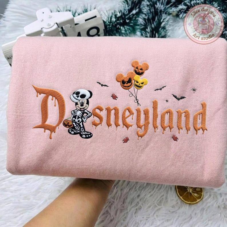 May include: A light pink sweatshirt featuring embroidered orange text that reads Disneyland in a dripping font. The design includes a skeleton-costumed mouse character, three jack-o'-lantern balloons, and small black bats scattered across the fabric.