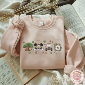 May include: A light pink sweatshirt with an embroidered design featuring a tree, binoculars, a jeep, and a compass. The design is in a row with green leaf accents. The sweatshirt has long sleeves and a round neckline.