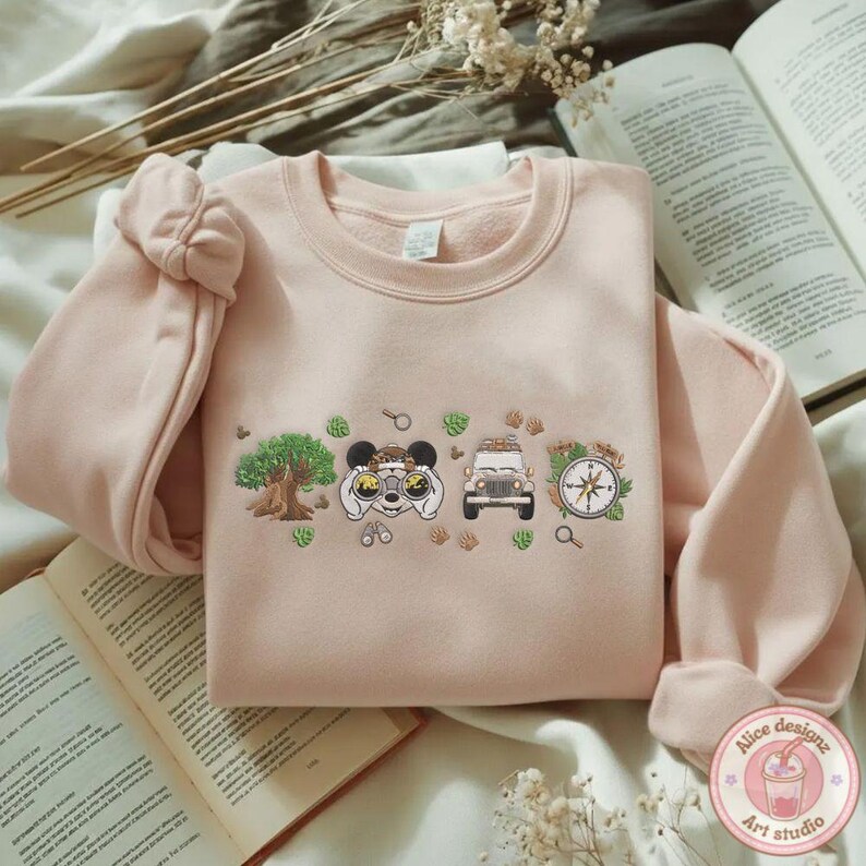 May include: A light pink sweatshirt with an embroidered design featuring a tree, binoculars, a jeep, and a compass. The design is in a row with green leaf accents. The sweatshirt has long sleeves and a round neckline.