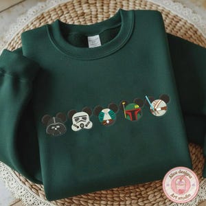 May include: Dark green sweatshirt featuring embroidered character designs. The design includes Darth Vader, a Stormtrooper, and other characters. The sweatshirt has a crew neck and long sleeves. The design is in the style of Mickey Mouse.