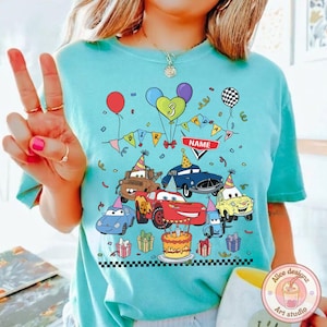 Op de afbeelding: Lichtblauw T-shirt met een kleurrijk verjaardagsontwerp met tekenfilmauto's, ballonnen en een taart. Het ontwerp bevat het nummer 3, een banner met "BIRTHDAY" en een ruimte voor een naam.