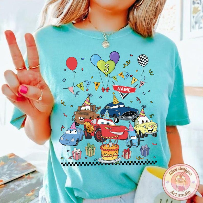 Op de afbeelding: Lichtblauw T-shirt met een kleurrijk verjaardagsontwerp met tekenfilmauto's, ballonnen en een taart. Het ontwerp bevat het nummer 3, een banner met "BIRTHDAY" en een ruimte voor een naam.