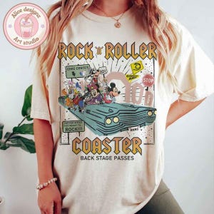 May include: A white t-shirt with a graphic print of cartoon characters riding in a vintage car. The text "Rock 'n' Roller Coaster Back Stage Passes" is printed on the shirt.