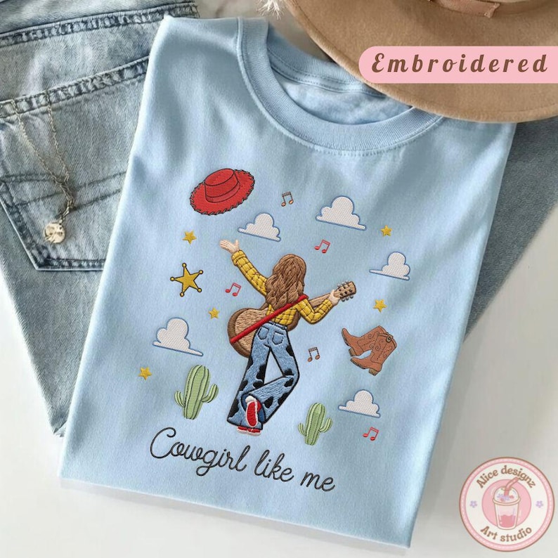 May include: A light blue t-shirt featuring an embroidered illustration of a person with long brown hair playing a guitar, wearing a yellow shirt and cow-print pants. The design includes a red hat, boots, cacti, stars, clouds, and the text Cowgirl like me.