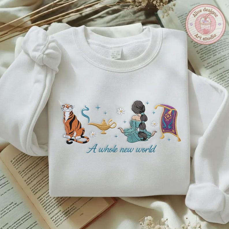 May include: A white crewneck sweatshirt featuring embroidered designs of a tiger, a gold lamp, a person with long dark hair in a teal dress, and a purple flying carpet. The text A whole new world is embroidered in blue below the illustrations.