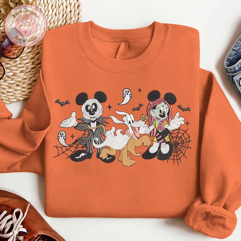 May include: Orange sweatshirt featuring a Halloween-themed graphic with cartoon characters. The design includes Mickey Mouse dressed as Jack Skellington, Pluto as a ghost, and Minnie Mouse as a zombie, with bats and spiderwebs.