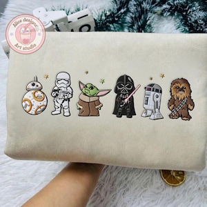 May include: Beige sweatshirt embroidered with Star Wars characters: BB-8, a Stormtrooper, Grogu, Darth Vader with a lightsaber, R2-D2, and Chewbacca. The design includes small star accents. The sweatshirt has a round logo in the upper left corner.
