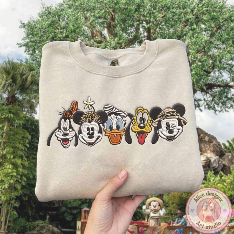 Puede incluir: Sudadera beige con caras de personajes de dibujos animados bordadas. Los personajes incluyen a Goofy, Mickey Mouse, Donald Duck, Pluto y un Mickey Mouse con tem&aacute;tica de safari. La sudadera es sostenida por la mano de una persona.