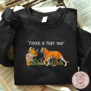 Op de afbeelding: Zwarte sweatshirt met een geborduurd ontwerp met Winnie de Poeh en Tijgertje. De tekst "Tigger, is that you?" staat boven de personages. Het ontwerp bevat een tijger en een beer in detective-outfit.