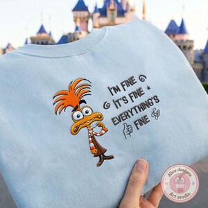 May include: Light blue sweatshirt featuring an embroidered cartoon character with orange hair and a brown outfit. The text "I'M FINE, IT'S FINE, EVERYTHING'S FINE" is embroidered on the front. A thumbs-up symbol is also embroidered.