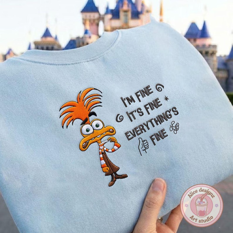 May include: Light blue sweatshirt featuring an embroidered cartoon character with orange hair and a brown outfit. The text "I'M FINE, IT'S FINE, EVERYTHING'S FINE" is embroidered on the front. A thumbs-up symbol is also embroidered.