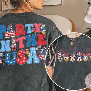 Peut inclure: T-shirt gris fonc&eacute; avec l'inscription "Party in the USA" en rouge, blanc et bleu, orn&eacute;e de personnages de dessins anim&eacute;s. Parfait pour c&eacute;l&eacute;brer le 4 juillet ou tout &eacute;v&eacute;nement patriotique.