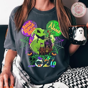 May include: Dark gray t-shirt featuring a Halloween-themed design. The graphic includes a green Oogie Boogie character, a purple castle, and the text "Boogie Bash". A black mug with a white ghost design is held in the hand.