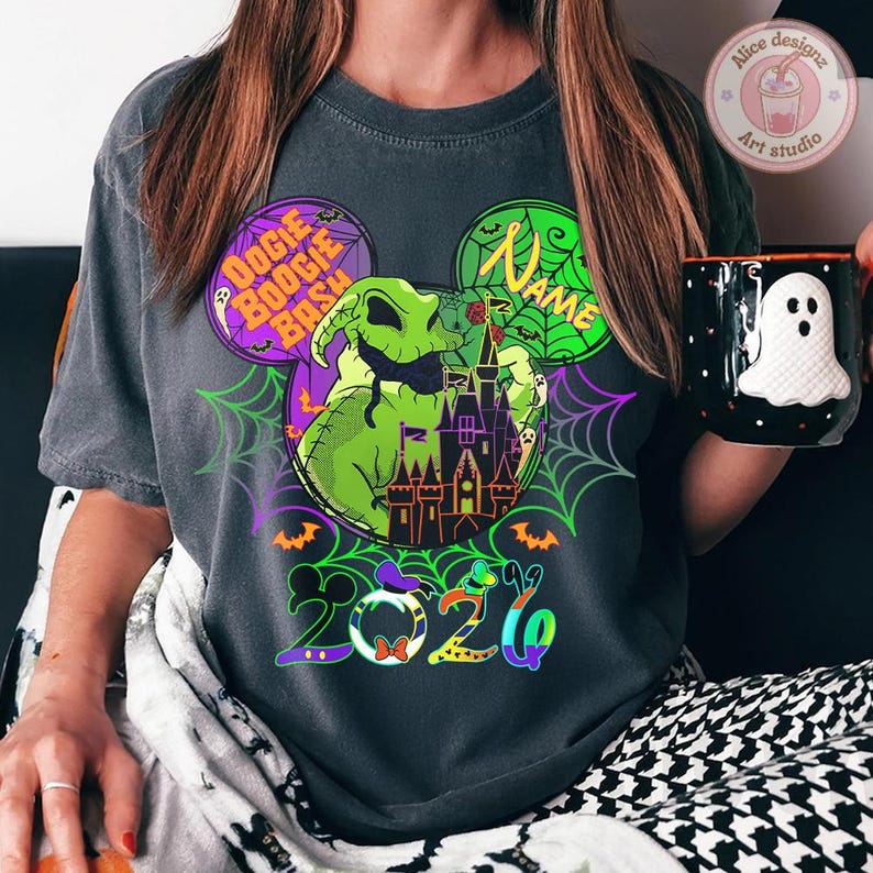 May include: Dark gray t-shirt featuring a Halloween-themed design. The graphic includes a green Oogie Boogie character, a purple castle, and the text "Boogie Bash". A black mug with a white ghost design is held in the hand.
