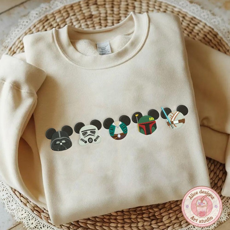 May include: Cream-colored sweatshirt featuring embroidered character designs. The designs include Darth Vader, a Stormtrooper, and other characters. The sweatshirt has a crew neck and long sleeves. The design is a mashup of Disney and Star Wars.