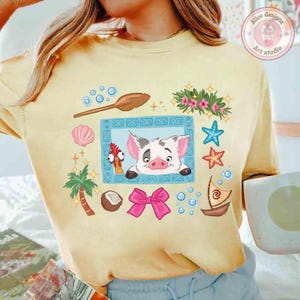 Puede incluir: Camiseta amarilla clara con una ilustraci&oacute;n de dibujos animados que presenta un cerdo, un pollo y elementos tropicales. El dise&ntilde;o incluye un lazo rosa, conchas marinas y un peque&ntilde;o barco. La camiseta tiene un corte holgado.