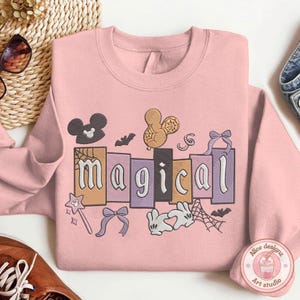 May include: Pale pink sweatshirt with the word "magical" in a playful font, decorated with Mickey Mouse ears, a bow, a magic wand, and other motifs. The design uses black, orange, purple, and white.