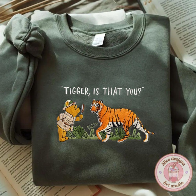 Op de afbeelding: Olijfgroene sweatshirt met een geborduurd ontwerp. Het ontwerp toont de tekst "Tigger, is that you?" boven een gele beer met een vergrootglas en een oranje en zwarte tijger. De sweatshirt heeft een ronde hals en lange mouwen.
