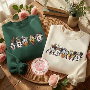 Puede incluir: Dos sudaderas de cuello redondo, una verde bosque y otra crema, cada una bordada con caras de personajes de dibujos animados. Las sudaderas se exhiben sobre una superficie de madera con detalles florales y una cesta de mimbre.