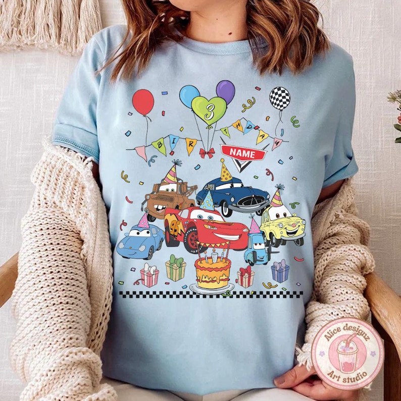 Op de afbeelding: Lichtblauw T-shirt met een kleurrijke print van tekenfilmauto's, ballonnen, confetti en een verjaardagstaart. Het ontwerp bevat het nummer "3" en het woord "NAME" op een banner. Het shirt heeft een ronde hals en korte mouwen.
