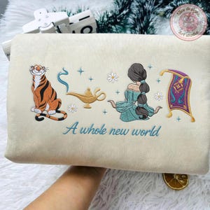 May include: A cream-colored sweatshirt featuring embroidered designs of a tiger, a gold lamp with blue smoke, a person with long dark hair in a teal outfit, and a purple magic carpet, with the text A whole new world stitched in blue thread.