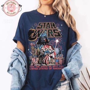 Op de afbeelding: Marineblauw T-shirt met een Star Wars-afbeelding met Darth Vader, vuurwerk en de tekst "MAY THE 250TH BE WITH YOU UNITED STATES OF AMERICA." Het ontwerp bevat Star Wars-personages en elementen van de Amerikaanse vlag.
