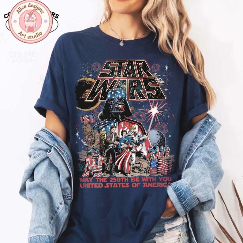 Op de afbeelding: Marineblauw T-shirt met een Star Wars-afbeelding met Darth Vader, vuurwerk en de tekst "MAY THE 250TH BE WITH YOU UNITED STATES OF AMERICA." Het ontwerp bevat Star Wars-personages en elementen van de Amerikaanse vlag.