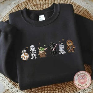 May include: Black crewneck sweatshirt featuring embroidered Star Wars characters: BB-8, a Stormtrooper, Grogu, Darth Vader, R2-D2, and Chewbacca. The design includes small gold stars. The sweatshirt is displayed on a woven surface.