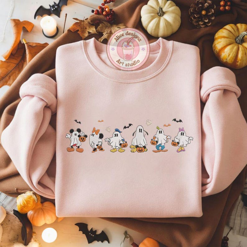 K&ouml;nnte beinhalten: Hellrosa Sweatshirt mit einem Halloween-Motiv. Die Grafik zeigt Zeichentrickfiguren in Geisterkost&uuml;men, die K&uuml;rbisse tragen. Das Sweatshirt hat lange &Auml;rmel und einen Rundhalsausschnitt. Das Bild enth&auml;lt auch K&uuml;rbisse und dekorative Flederm&auml;use.