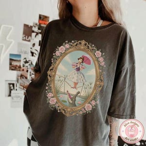 May include: A charcoal gray short-sleeve t-shirt featuring a central graphic of a person in a dress holding a pink umbrella while walking on a tightrope above an alligator. The scene is enclosed in an ornate gold oval frame with pink roses.