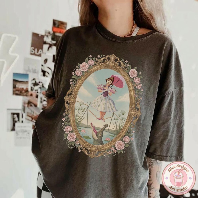 May include: A charcoal gray short-sleeve t-shirt featuring a central graphic of a person in a dress holding a pink umbrella while walking on a tightrope above an alligator. The scene is enclosed in an ornate gold oval frame with pink roses.
