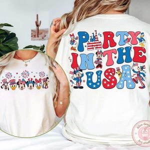 Peut inclure: T-shirt blanc cass&eacute; avec un motif graphique color&eacute;. Le devant pr&eacute;sente des personnages de dessins anim&eacute;s avec des chapeaux patriotiques et des feux d'artifice. Le dos du t-shirt dit "Party in the USA" en lettres rouges et bleues.
