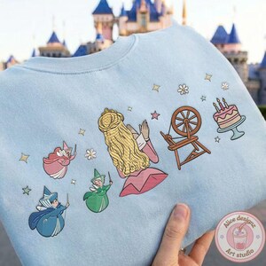 May include: Light blue sweatshirt featuring an embroidered design of a princess, fairies, a spinning wheel, and a cake. The design includes stars and flowers. The sweatshirt is a soft, comfortable material.