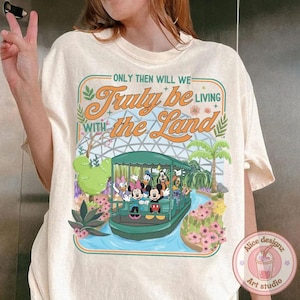 May include: Cream-colored t-shirt featuring a graphic of a boat with Disney characters, including Mickey and Minnie Mouse, cruising through a garden. The text "ONLY THEN WILL WE Truly be LIVING WITH the Land" is above the image. A small logo for "Alice designz Art studio" is in the bottom right.