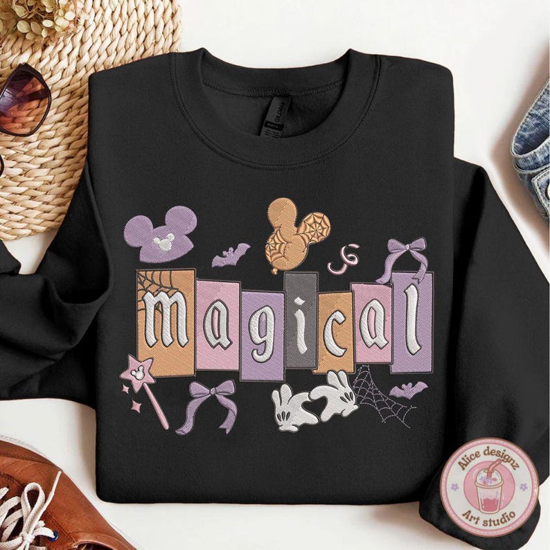 May include: Black crew neck sweatshirt featuring the word "magical" in a colourful, block-letter design. The design includes Mickey Mouse silhouettes, bats, bows, a wand, and a spiderweb.