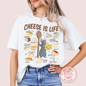 Puede incluir: Camiseta blanca con la frase "CHEESE IS LIFE" e ilustraciones de quesos. Un chef rata de dibujos animados sostiene una cuchara de madera y un bloque de queso. Las variedades de queso incluyen Gouda, Brie y Camembert.