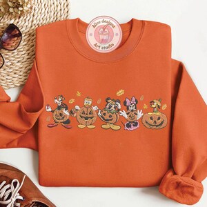 Puede incluir: Sudadera naranja con una fila de personajes de dibujos animados dentro de calabazas talladas. Los personajes incluyen un perro, el Pato Donald, Mickey Mouse, Minnie Mouse y otro perro. La sudadera tiene el texto "Alice designz Art studio".